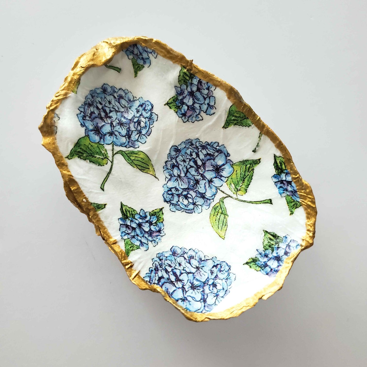 Oyster shell jewelry dish with a blue hydrangea design on a white background and gold painted edges