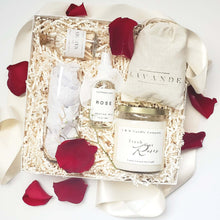Load image into Gallery viewer, A gift box for a bride or bride-to-be which includes a glass stemless champagne flute with the word Bride engraved on it in gold, a premium soy candle with the scent of fresh cut roses, white tip matches in a apothecary-inspired glass bottle with a cork top, rose water hydrating facial mist and a muslin lavender sachet bag with a drawstring and real red rose petals sprinkled throughout.
