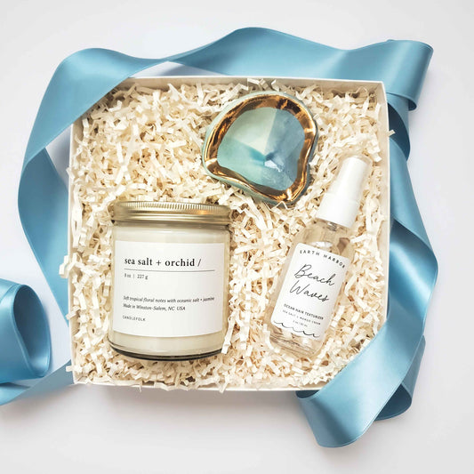 A beach inspired curated gift box that includes a hand poured sea salt and orchid candle that has soft floral notes with oceanic salt and jasmine. Also included is a beach waves ocean hair texturizer and a hand made blue toned jewelry dish with edges painted with 22k gold. 