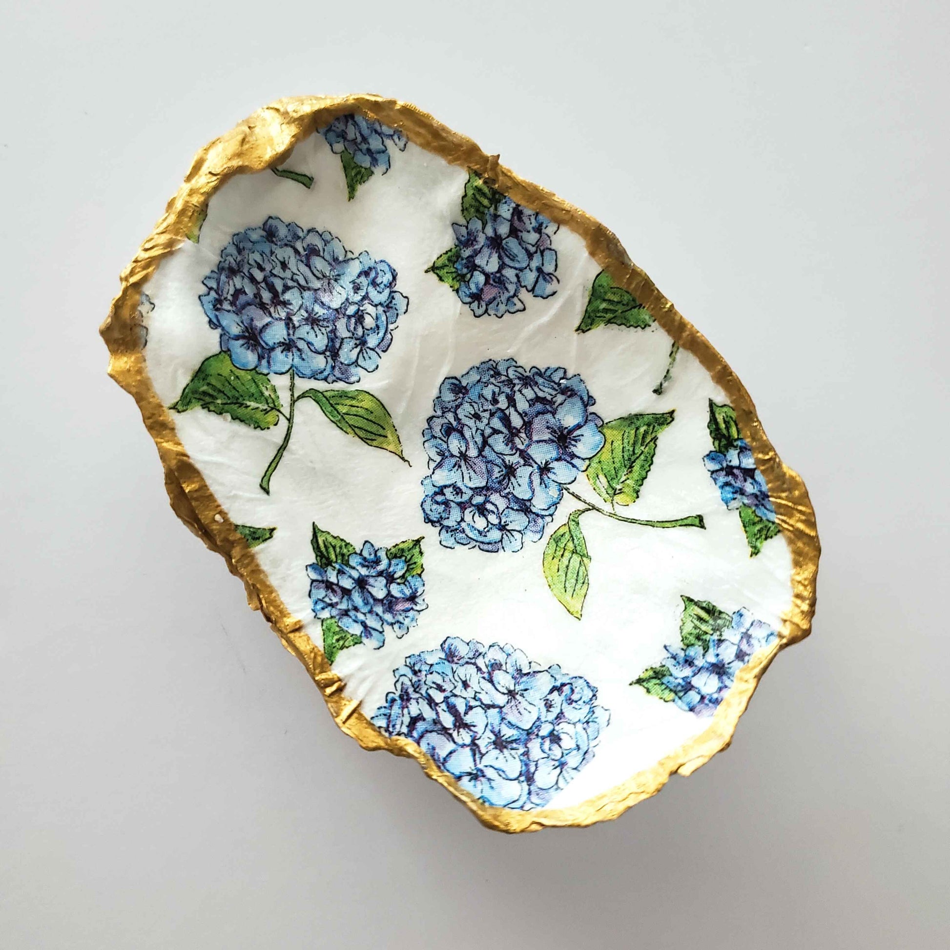 Oyster shell jewelry dish with a blue hydrangea design on a white background and gold painted edges