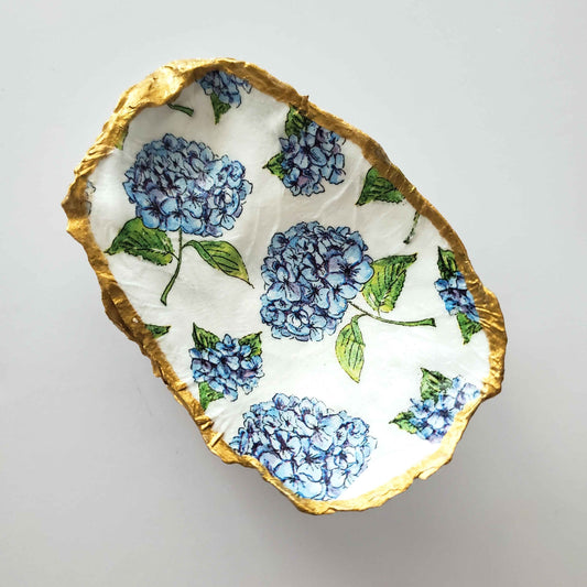 Oyster shell jewelry dish with a blue hydrangea design on a white background and gold painted edges