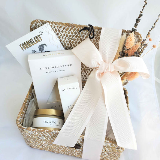 A spa like gift set which includes a travel size candle, luxe headband, tidal rose mist and Formulary 55 facial mask all packaged in a seagrass lidded storage basket with dried florals and a light pink ribbon.