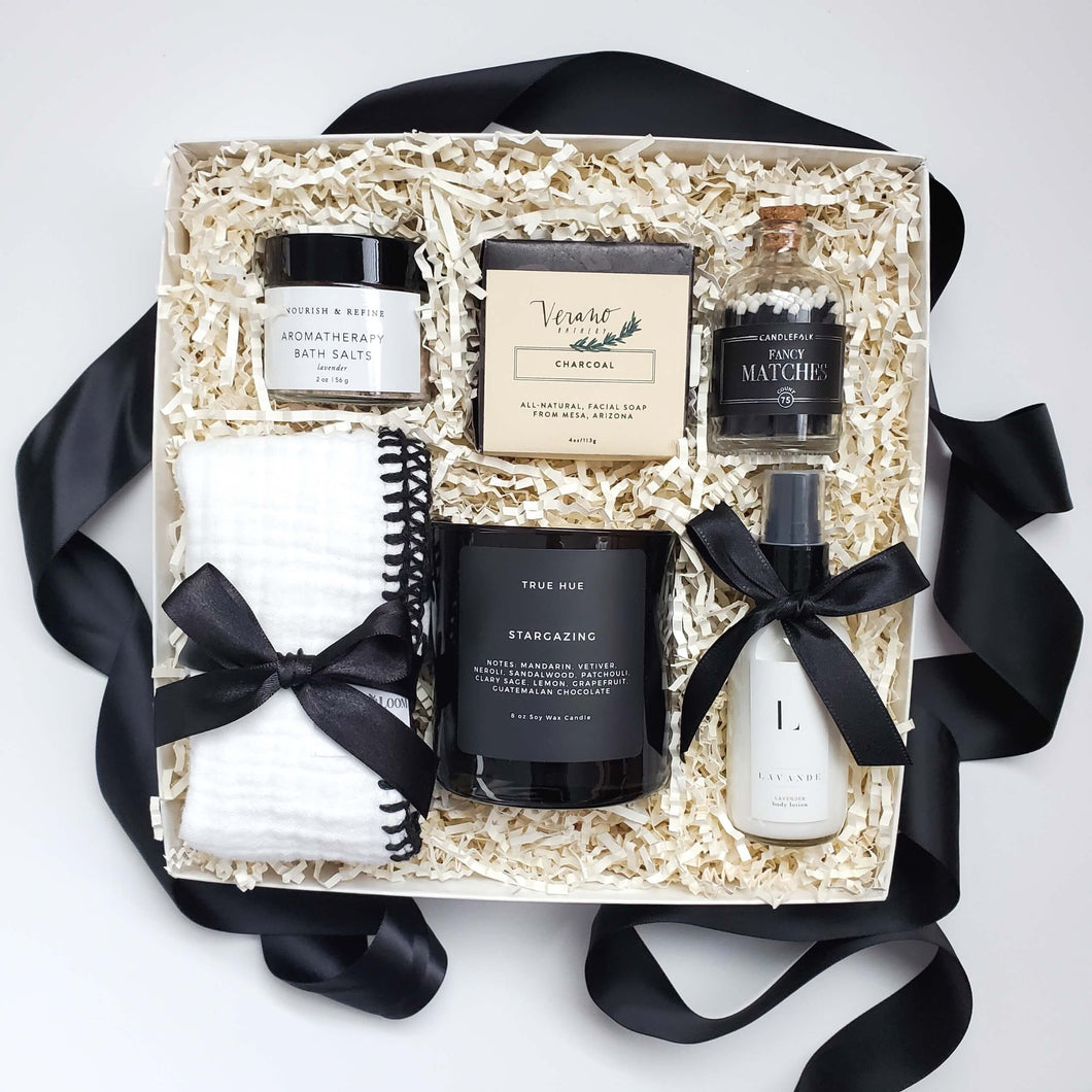 A spa gift box with a black and white color scheme which includes  charcoal facial soap, face towel, aromatherapy bath salts, moisturizing lotion, a soy candle and black and white matches in a apothecary-inspired bottle.