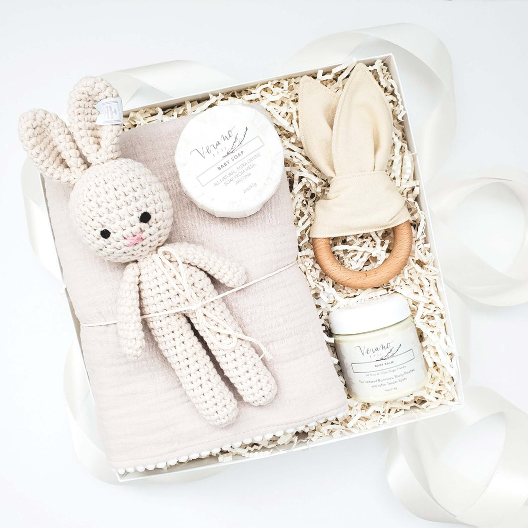 Gender neutral baby gift box from The Gift Veil featuring a toy bunny rattle, bunny ears teether, muslin swaddle blanket, gentle baby soap and baby balm.