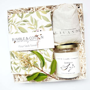 A housewarming gift box by The Gift Veil featuring a hand-poured juniper and sage candle, a flour sack towel with green spring leaves print and a lavender sachet.