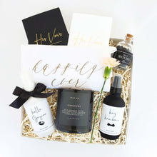 Load image into Gallery viewer, An engagement or wedding gift box with a black and white color palette by The Gift Veil. Includes his and her vow books, a soy candle, black matches, mood sprays and a tea towel that says happily ever after.
