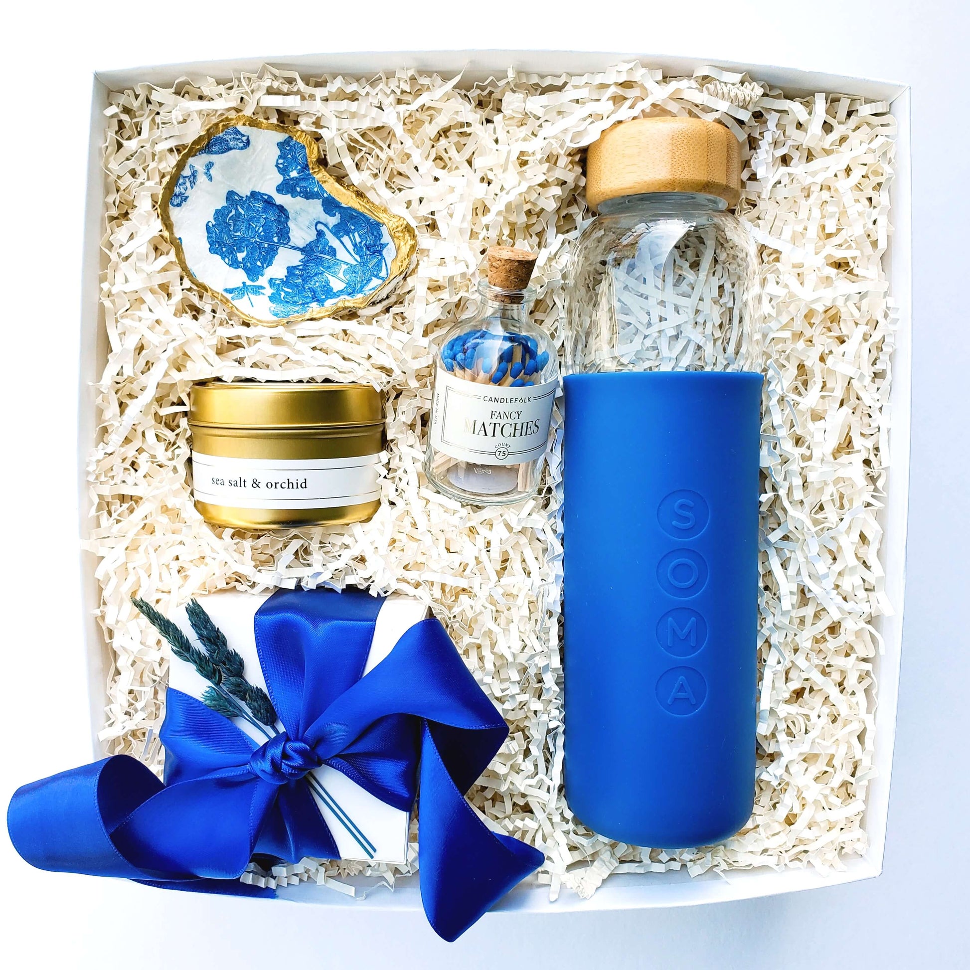 A curated gift box which includes a blue glass water bottle, a bluebird oyster shell jewelry dish, matches in a glass apothecary bottle, a travel size candle, and a small treat box of sea salt caramels. 